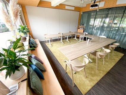 Vergaderruimte bij Napkins Woerden met moderne houten tafels, beige stoelen en grote ramen die veel daglicht binnenlaten. De ruimte is sfeervol ingericht met groene planten, kussens op de vensterbank en een zachtgroen vloerkleed, ideaal voor een inspirerende meeting of workshop.