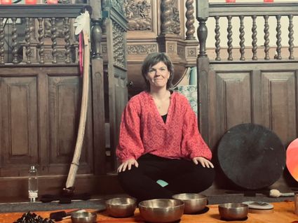 Op de foto is Daphne Posthuma van Praktijk Wad 'n Klank te zien. Ze zit tussen haar klankschalen en andere instrumenten zoals een drum, rainstick en een sansula. Ze is er klaar voor om haar klankschaalconcert te starten.