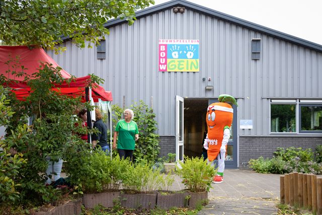 Op de achtergrond het hoofdgebouw van Bouwgein, daarvoor de tuinen en vrijwilligers. Met Joggy, de mascotte van de JOGG aanpak.