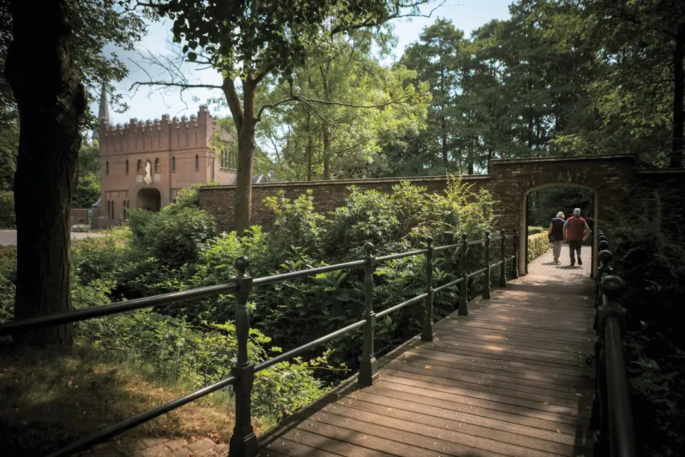 Een stel loopt over het bruggetje naar het terras en horeca van de Abdij. Deze zie je in de verte liggen in het groen