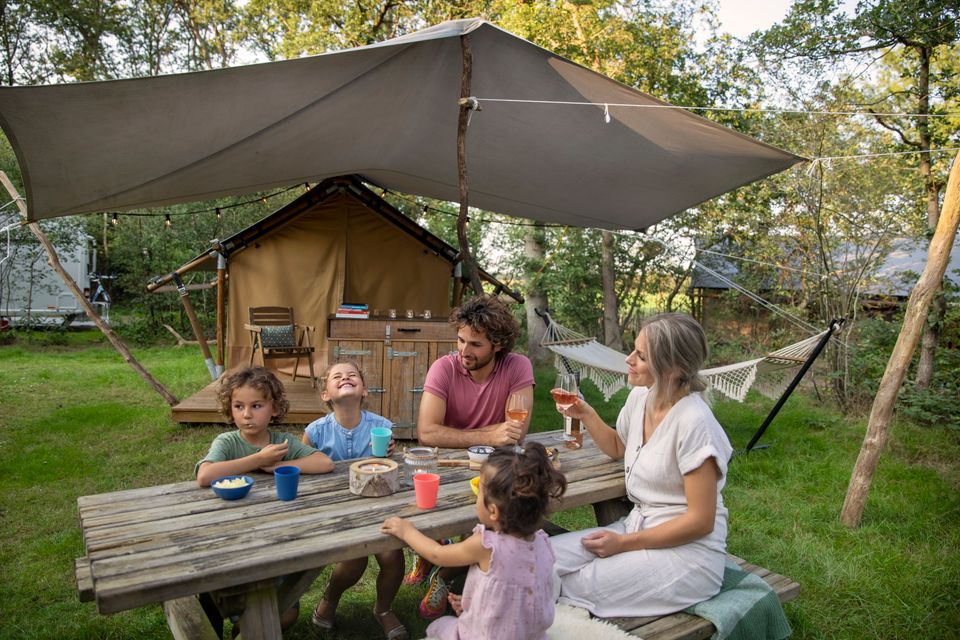 Een gezin zit gezellig aan de picknicktafel voor hun safaritent.