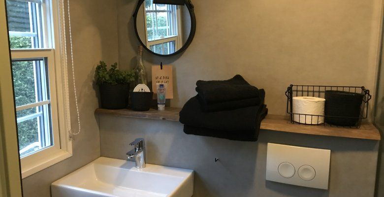 Detailfoto van een wastafel met  houten plank daarboven met handdoekjes, mandje met toiletpapierrollen en een ronde spiegel erboven.