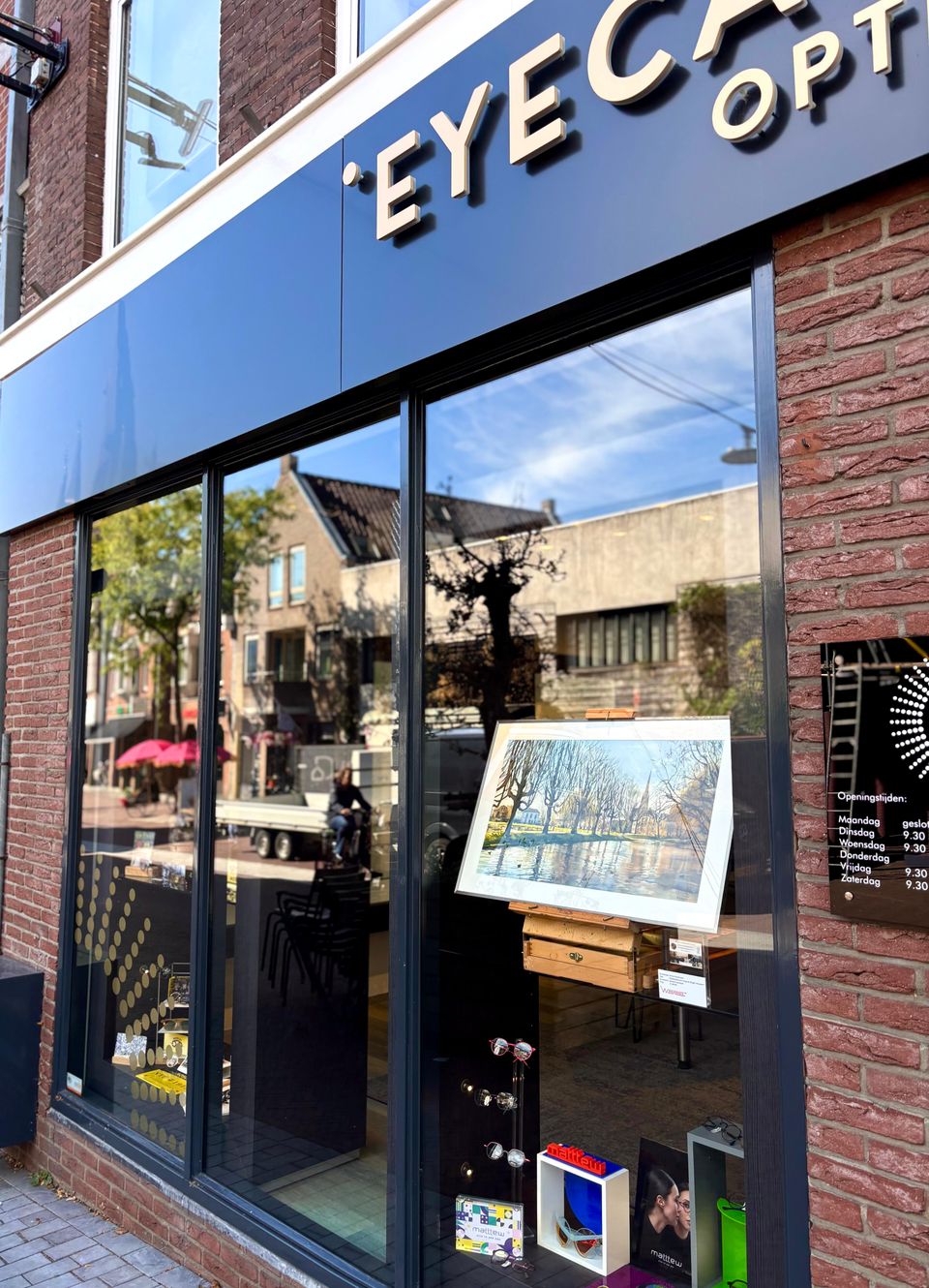 Winkelpand van EyeCare Optiek in Woerden, Groene Hart, glazen pui met reflectie van de binnenstad en etalage met ingelijst schilderij op ezel en diverse monturen tijdens de Atelierroute Woerden.