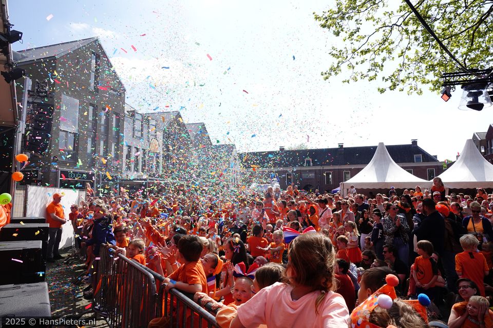 Publieksevenement Koningsdag in Woerden, Groene Hart, groot kinderfeest met confetti, podium en feestende gezinnen in oranje kleding op een plein in de historische binnenstad.