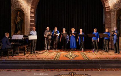Overzicht van het kleinkoor tijdens een optreden. De leden dragen blauwe en zwarte kleding.