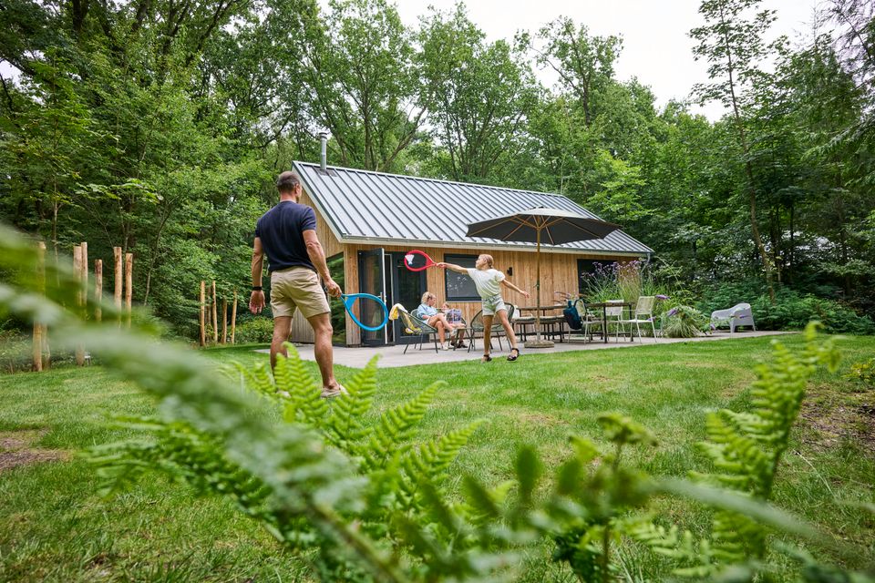 Vader en dochter aan het badmintonnen in de tuin bij Boshuis Bambi in Oudemirdum terwijl moeder en dochter op de achtergrond in het inspiratiemagazine van VVV Waterland van Friesland bladeren.