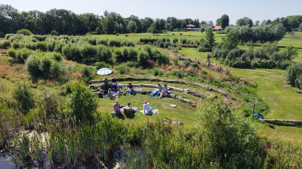 Groep mensen zit in het natuurlijke amfitheater van Buitenplaats Kameryck, omringd door riet, struiken en weidse natuur.