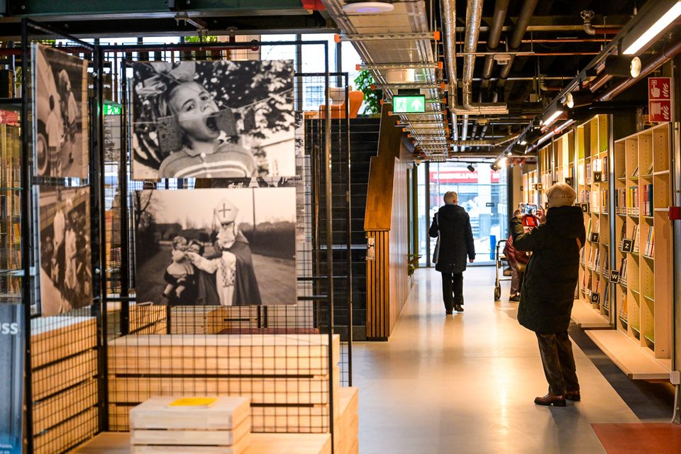 Interieur van een moderne bibliotheek met een fototentoonstelling: zwart-witfoto’s hangen in rekken, langs de boekenkasten lopen bezoekers door de gang, één persoon maakt een foto met een telefoon.