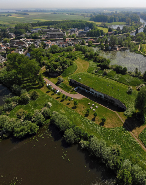 Een bakstenen fort overgroeid met planten en bomen, omgeven door een fortgracht en daarachter een klein dorp.
