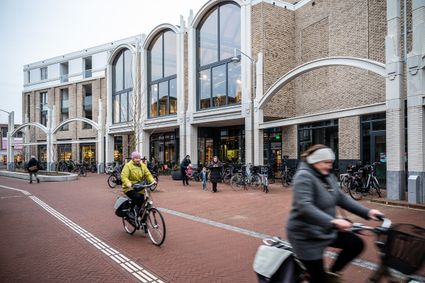 Straatbeeld met fietsers en voetgangers voor een modern bakstenen gebouw met grote boogramen, waar mensen binnenkomen en fietsen geparkeerd staan bij de ingang.