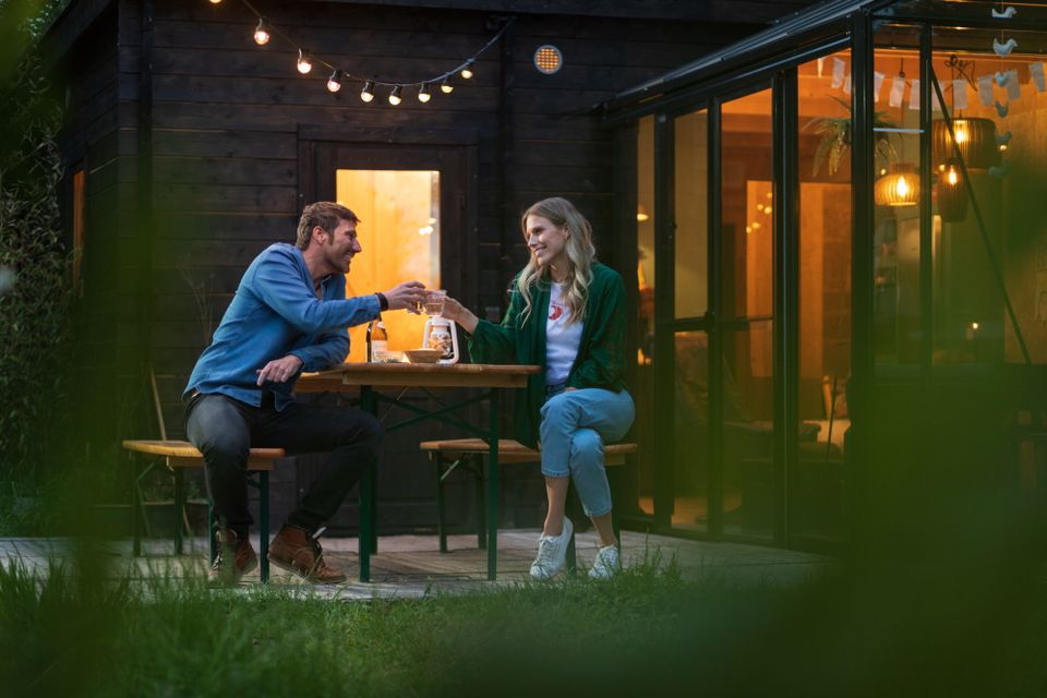 Een man en vrouw zitten buiten op een bankje bij hun vakantiehuis en proosten lachend met elkaar.