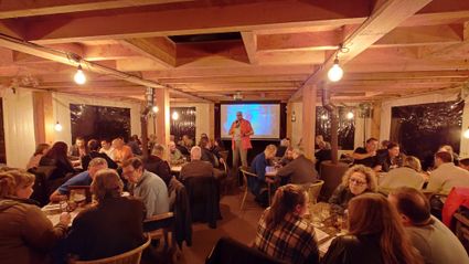 Foto van het interieur van De Rode Lelie met diverse zitjes met gasten. De quiz-master staat in het midden voor een groot beeldscherm.