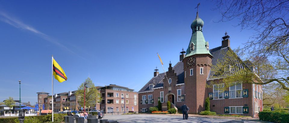 Gemeentehuis van Woudenberg aan het centrale plein, met vlaggen, zitgelegenheid en bebouwing van het dorpscentrum op de achtergrond. De foto laat het karakteristieke raadhuis en de centrale ligging in Woudenberg zien.