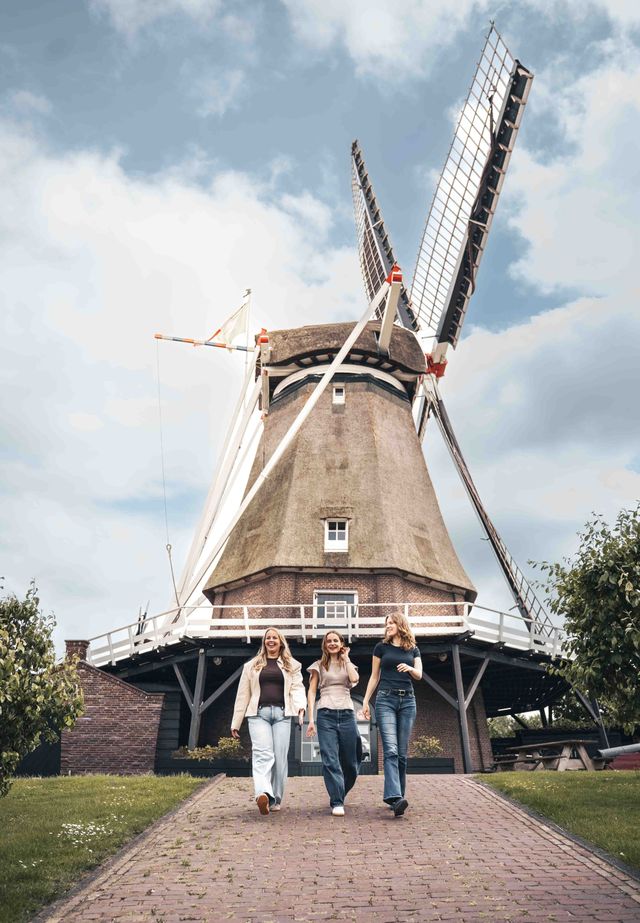 Korenmolen De Valk in Montfoort, Groene Hart. Drie bezoekers wandelen over het pad voor de historische windmolen met rieten romp en draaiende wieken onder een bewolkte Hollandse lucht.