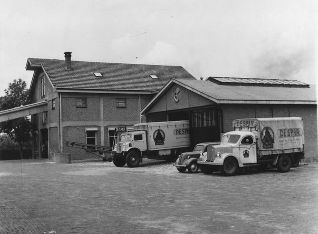 Oude zwart/wit foto van het pakhuis van de Spar aan de Dorpsstraat 180, met 2 vrachtwagens ervoor.