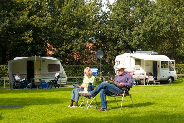 Foto van een groen grasveld met aan de rand bomen, waaronder 2 caravans staan en 2 gasten op hun campingstoeltjes op de voorgrond.