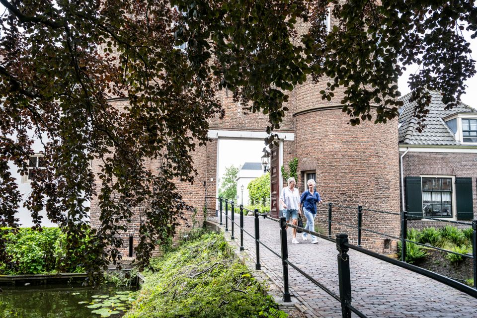 Twee wandelaars lopen over een brug langs het water bij Kasteel Montfoort, met op de achtergrond de ronde bakstenen torens en het groen van de historische vesting.