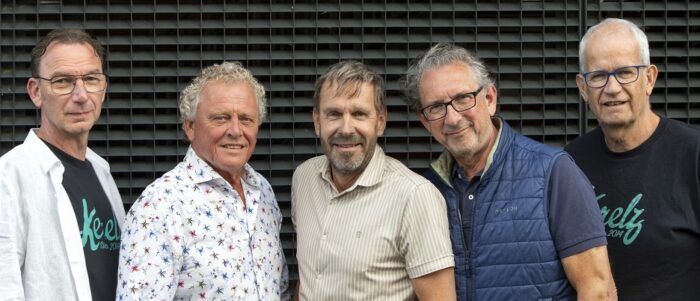 Vijf volwassen mannen staan naast elkaar en kijken in de camera. Ze dragen casual kleding en staan voor een metalen gevel. De foto oogt als een informeel groepsportret, mogelijk van collega’s of bestuursleden.