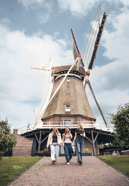 Historische korenmolen De Valk in Montfoort, Groene Hart, provincie Utrecht. Drie vrouwen wandelen over het klinkerpad voor de traditionele Nederlandse windmolen met rieten kap en grote wieken.