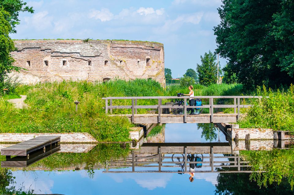 Een vrouw staat op een brug samen met haar fiets. Op de achtergrond is een rond fort zichtbaar.