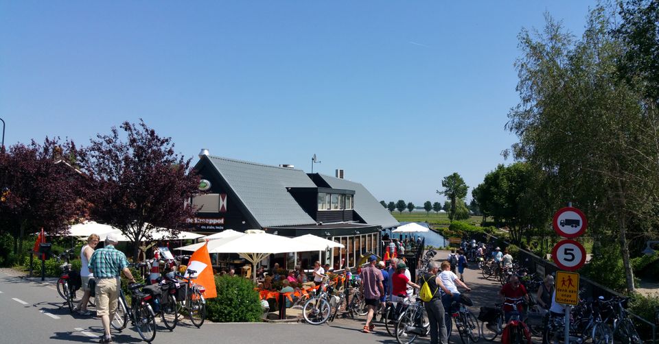 Restaurant De Strooppot in De Hoef, Groene Hart. Druk terras aan het water met fietsers, geparkeerde fietsen en bezoekers tijdens een zonnige fietstocht door de regio.