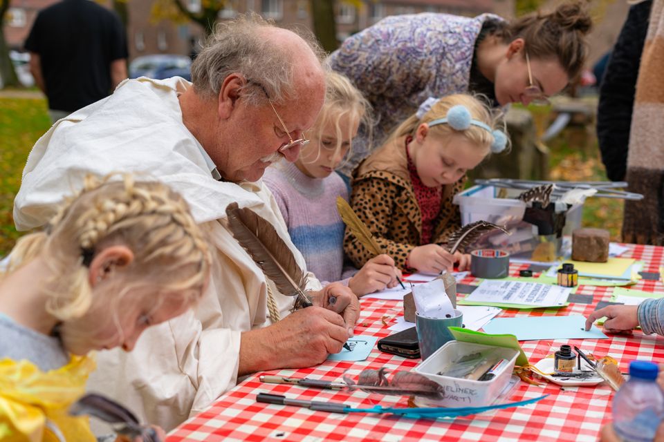 Man en kinderen die samen met ganzenveren en inkt schrijven aan een tafel met rood-wit geruit tafelkleed tijdens een creatieve workshop buiten.