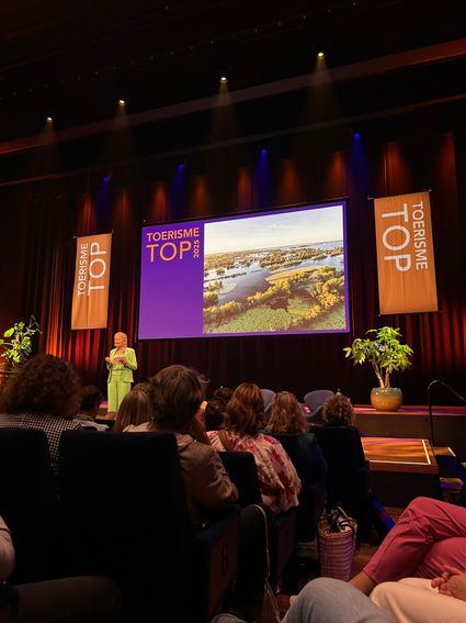 Een zaal vol mensen, kijkend naar een vrouw in een groen pak die op het podium staat te presenteren, met op de achtergrond een enorm scherm waarop Toerisme TOP 2025 staat.