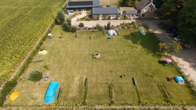Luchtfoto van een kleinschalig kampeerterrein met tenten en picknicktafels op een grasveld naast een boerderij en maïsveld.