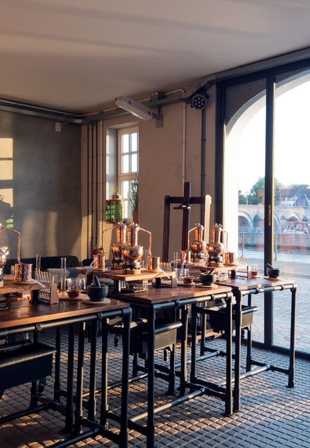 Ambachtelijke ginworkshop bij Het Stookatelier in Maastricht met uitzicht op de Maas. De koperen distilleerketels staan klaar op houten werkbanken in een sfeervolle ruimte aan de Oeverwal 8.