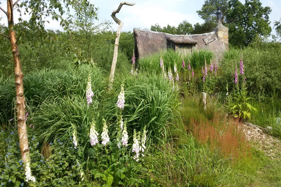 Een plaggenhut omringd door bomen, struiken en paarse bloemen.