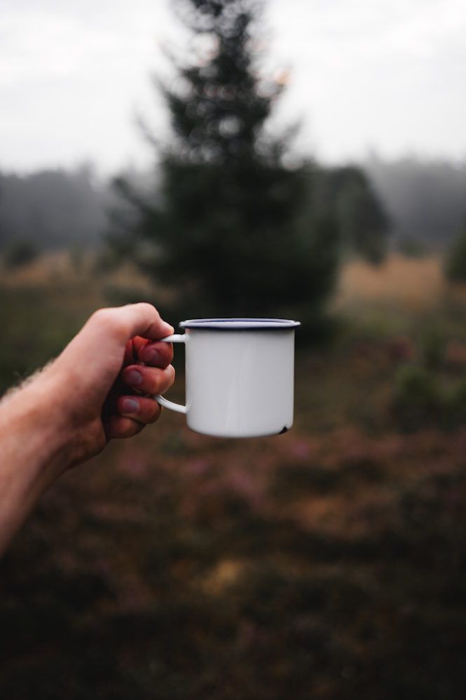 Een hand houdt een witte emaille mok vast. Op de achtergrond zie je de natuur in Drenthe.