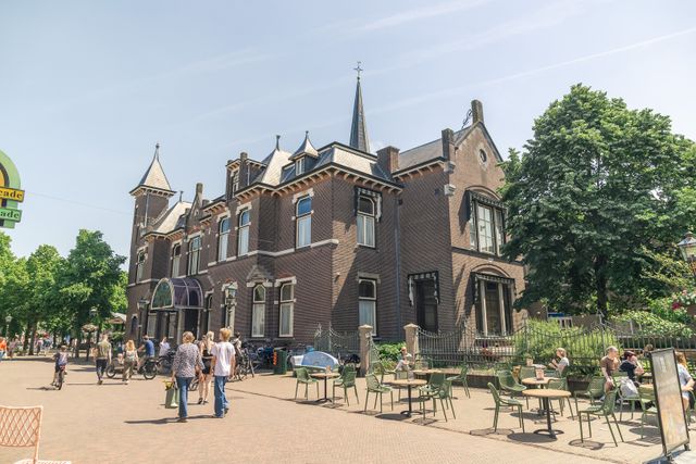 Zonnige dorpskern van Baarn met een historisch pand, terrassen en bezoekers op de Brink. De foto laat de levendige en groene sfeer van het centrum van Baarn zien.