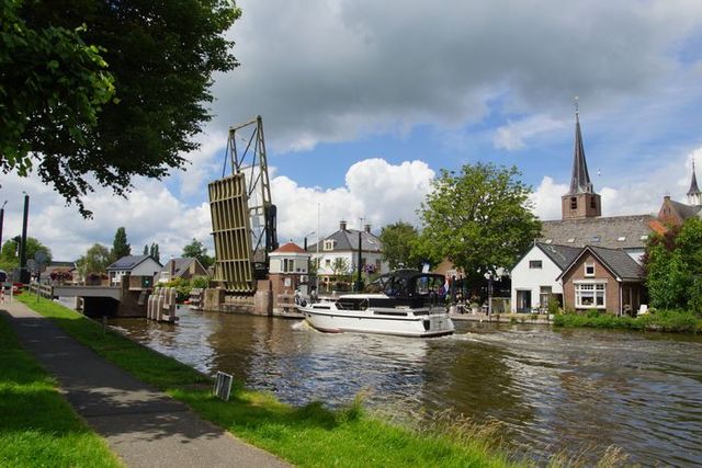 Pleziervaartuig vaart onder een openstaande ophaalbrug door in Alphen aan den Rijn, met zicht op kerktoren, dorpshuizen en een bewolkte zomerlucht.