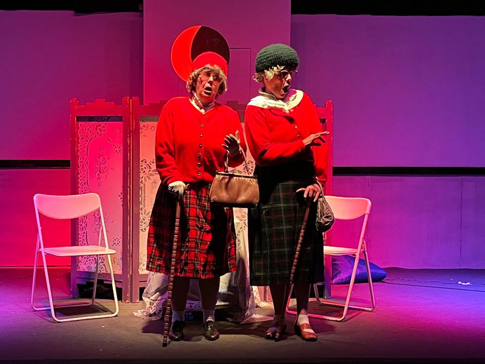 Foto van 2 oude dames met mutsen op, rode vestjes en schotse rokken aan tegen een rood-paarse achtergrond met 2 stoeltjes en een kamerscherm.