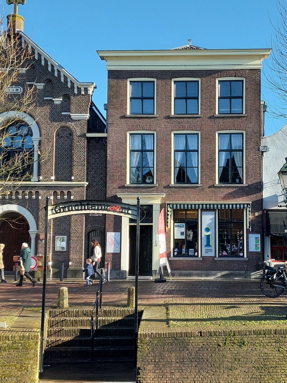 Toeristisch informatiepunt TIP Oudewater, Groene Hart. Historisch grachtenpand aan de gracht met bezoekersinformatie, naast een monumentale kerk en brug, terwijl wandelaars passeren in de binnenstad op een zonnige dag.