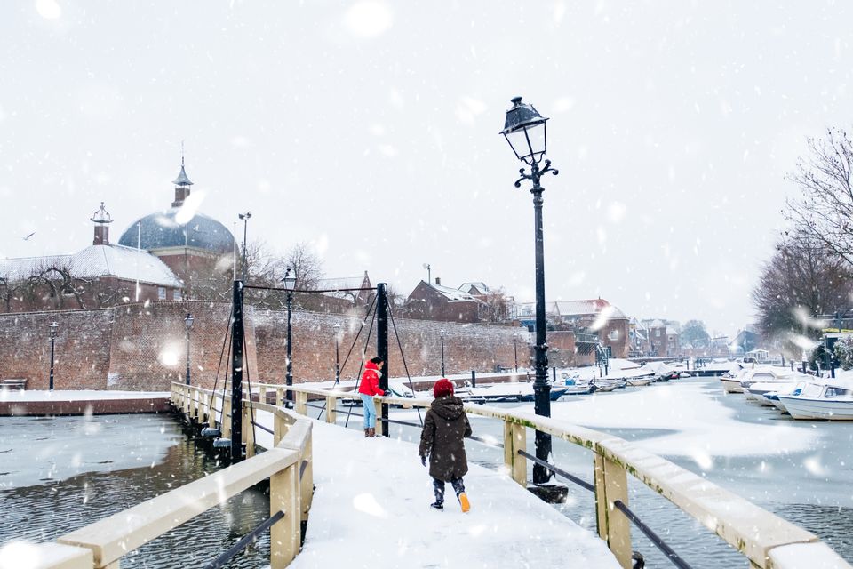 Twee kinderen lopen over een besneeuwde brug in vestingstad Leerdam, met op de achtergrond historische muren, boten aan de kade en vallende sneeuwvlokken – winterse sfeer in het Groene Hart.