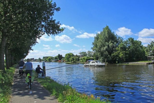 Fietsers en vissers genieten van het mooie weer langs de Oude Rijn in Bodegraven-Reeuwijk, terwijl een pleziervaartuig voorbij vaart onder een blauwe lucht.