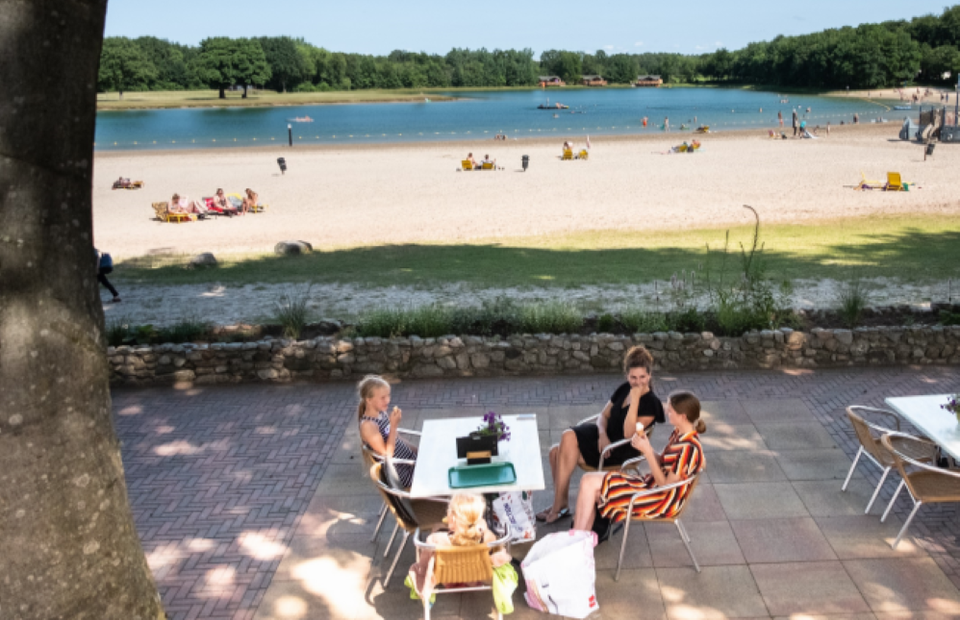 Eine Familie auf der Terrasse bei Ronostrand. Im Hintergrund liegt der See mit dem Strand.
