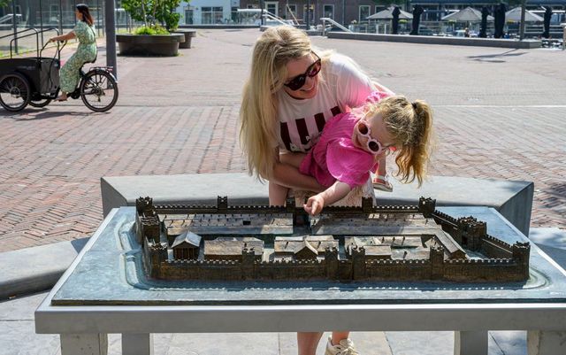 Een jonge vrouw met meisje op de arm buigen zich over de bronzen maquette van het fort Albaniana.
