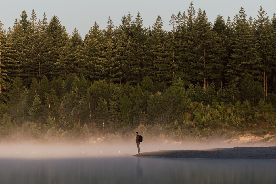 Ein Mann steht in der Ferne am Wasser, über dem ein leichter Nebel hängt.