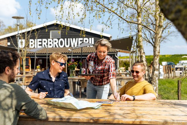 Verschillende mensen aan tafel op het terras bij Brouwerij Ooijen die een drankje geserveerd krijgen.
