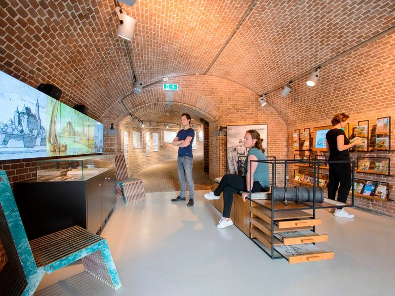 Een man en twee vrouwen staan en zitten in een bakstenen ruimte. Een vrouw en de man kijken naar een scherm waarop een animatie met oude prenten wordt afgespeeld. De tweede vrouw bestudeert folders in een meubel.