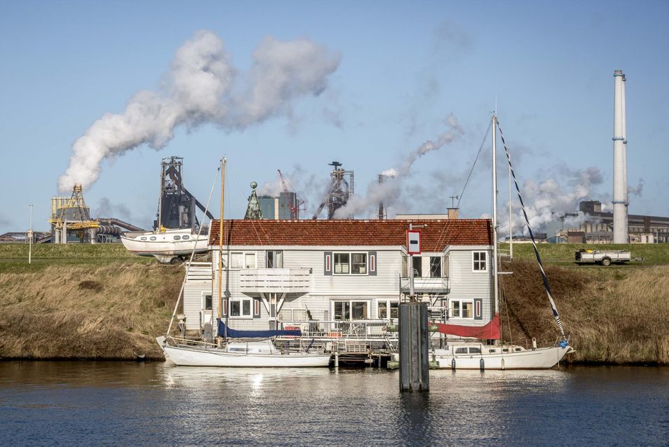 Industrieel landschap woning aan het water, woonhuis met aangemeerde schepen, Groene Hart, contrast tussen rustige waterkant en industriële installaties met rookpluimen op de achtergrond.