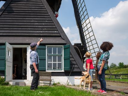 Drie personen staan bij een molen, de molenaar wijst omhoog.