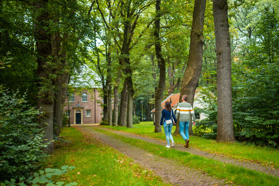 Een man en een vrouw wandelen over een pad omringt door groene bomen richting een ronde kerk (Koepelkerk) in Veenhuizen.