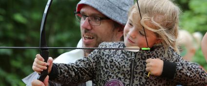Volwassene helpt een jong kind bij het boogschieten in een groene buitenomgeving. Het kind houdt de boog vast en richt een pijl, terwijl de volwassene achter hen meekijkt en ondersteunt.