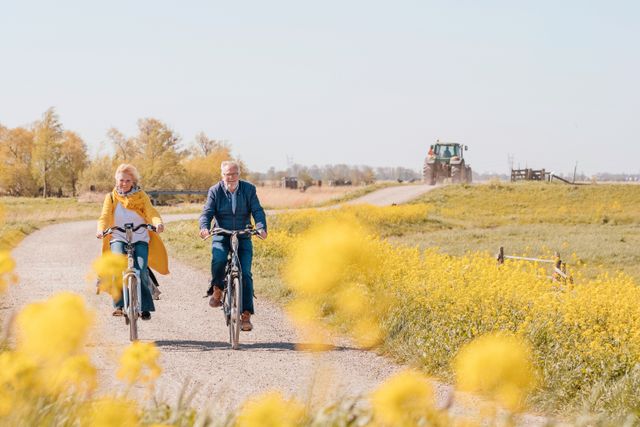 Twee fietsende mensen door de polder, met gele bloemetjes in de voorgrond en op de achtergrond zie je en tractor.