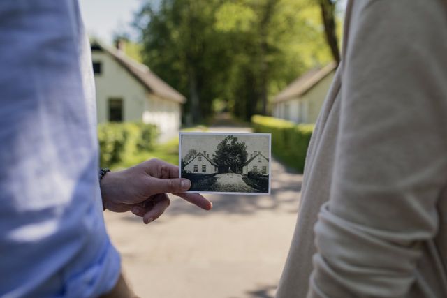 Op de voorgrond wordt een oude foto vastgehouden van koloniehuisjes die je op de achtergrond ziet.