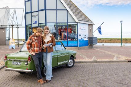 Twee mensen bekijken een informatiefolder van de VVV terwijl ze voor de VVV winkel in Stavoren staan, leunend tegen een auto.