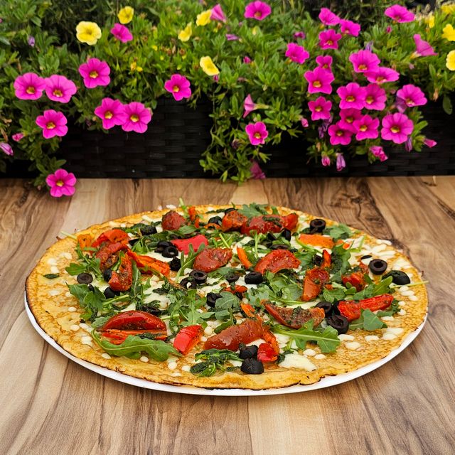 Pannenkoekenrestaurant De Strooppot in De Hoef, Groene Hart. Hartige pannenkoek met rucola, tomaat, paprika en zwarte olijven op een houten tafel met kleurrijke bloemen.